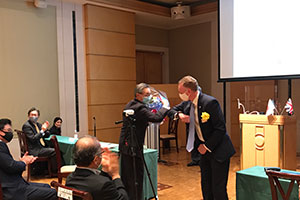 当日誕生日だった朝田会長にクリス・へファー駐日英国大使館公使参事官より花束贈呈、固い“コロナ握手”。
